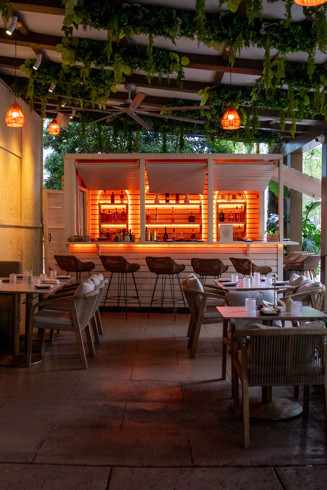 The plant-covered ZIVA patio with a glowing wood bar, rattan stools and set tables